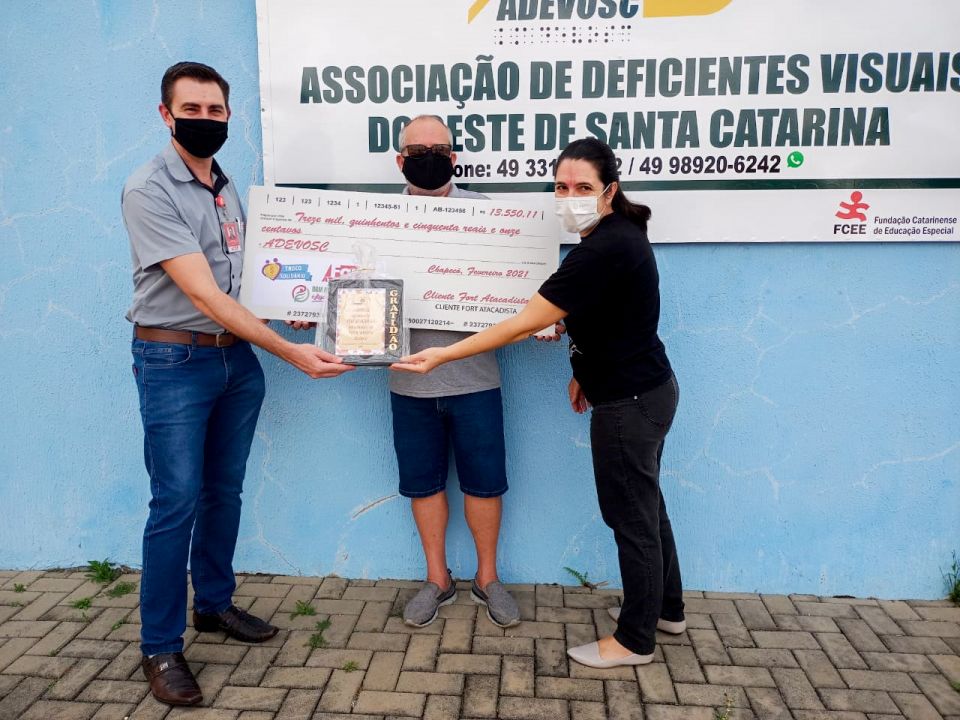 MB Comunicao Empresarial e Organizacional - #PARATODOSVEREM: FOTOGRAFIA DIANTE DA ADEVOSC, DO SR. DERSON FIORESE (GERENTE DO FORT ATACADISTA), COM O SR. ANTONIO CARLOS RIGO, PRESIDENTE DA ADEVOSC, E A SR. MIRIAM STEFFEN, DIRETORA ADMINISTRATIVA DA ADEVOSC. TODOS USANDO MSCARA E SEGURANDO CHEQUE REPRESENTATIVO E NOMINAL PARA A ADEVOSC, BEM COMO QUADRO COM A PALAVRA GRATIDO REPASSADO DA ADEVOSC PARA O FORT ATACADISTA. AO FUNDO DA IMAGEM ENCONTRA-SE PAINEL COM LOGO DA ADEVOSC.